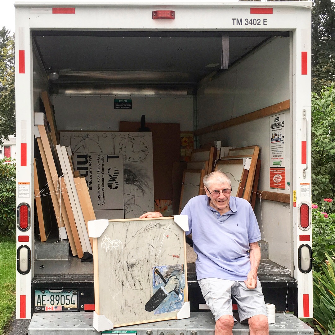 Robert Cartmell holding a self-portrait painting while sitting on the back of a U-Haul filled with his artwork during a Studio Visit. Photo credit: Anna Wettergreen